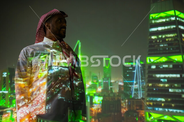 Distinctive architectural engineering art, a young Saudi Gulf Arab man wearing a traditional dress standing in front of towers and skyscrapers illuminated in green at night in the city of Riyadh on Saudi National Day, September 23, growth, progress and urban development in the Kingdom of Saudi Arabia, famous tourist attractions