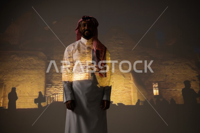 الأماكن القديمة الأثرية، المباني والبيوت الطينية المضاءة في وقت الليل، ايماءات السعادة والبهجة، شاب عربي خليجي سعودي مبتسم يرتدي الثوب التقليدي والشماغ يقف أمام أسوار قصر سلوى، زيارة المناطق السياحية في المملكة العربية السعودية