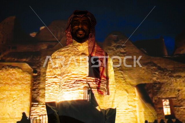 Gestures of happiness and joy, mud buildings and houses illuminated at night, ancient archaeological sites, a smiling Saudi Gulf Arab young man wearing the traditional dress and shemagh standing in front of the walls of Salwa Palace, visiting tourist areas in the Kingdom of Saudi Arabia