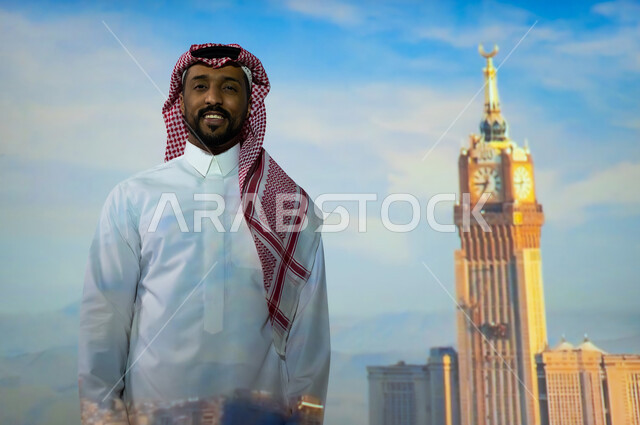 الأبراج وناطحات السحاب في المملكة العربية السعودية، المعالم الاسلامية الدينية المقدسة في السعودية, الفن الهندسي المعماري للمنشآت والمباني، شاب عربي خليجي سعودي يرتدي الثوب التقليدي يقف أمام برج الساعة الملكي في مكة المكرمة في وضح النهار ينظر إلى الكاميرا، ايماءات السعادة والبهجة