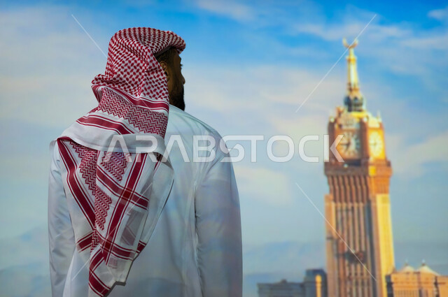 الفن الهندسي المعماري للمنشآت والمباني، المعالم الاسلامية الدينية المقدسة في السعودية, شاب عربي خليجي سعودي يرتدي الثوب التقليدي يقف أمام برج الساعة الملكي في مكة المكرمة في وضح النهار، الأبراج وناطحات السحاب في المملكة العربية السعودية