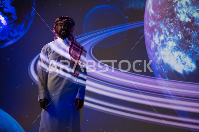 نمو قطاع الفضاء في المملكة العربية السعودية، دمج العلم بالتكنولوجيا، رؤية مستقبلية سعودية ٢٠٣٠، التطور الرقمي والتكنولوجي، شاب عربي سعودي خليجي يرتدي الثوب التقليدي يقف أمام الكواكب والنجوم وينظر إلى الكاميرا، خلفية هيولوجرامية ثلاثية الأبعاد للكواكب