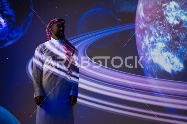 الاهتمام بقطاع الفضاء في المملكة العربية السعودية، دمج العلم بالتكنولوجيا، رؤية مستقبلية سعودية ٢٠٣٠، التطور الرقمي والتكنولوجي، شاب عربي سعودي خليجي يرتدي الثوب التقليدي يقف أمام الكواكب والنجوم، خلفية هيولوجرامية ثلاثية الأبعاد للكواكب