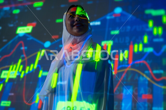 مفهوم التحليل المالي وفرص الاستثمار, سوق الأوراق المالية, امرأة عربية خليجية سعودية محجبة تقف أمام شاشة مليئة بالأرقام و الإحصائيات, صعود و هبوط أسهم البورصة , خلفية هولوجرامية ثلاثية الأبعاد