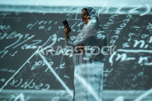 أرقام وحسابات في مادة الرياضيات، استخدام التقنيات الحديثة, دمج العلم بالتكنولوجيا، إمرأة عربية خليجية سعودية مبتسمة محجبة ترتدي العباءة تمسك بيدها الهاتف المحمول, التطور العلمي و التقني في المملكة العربية السعودية, خلفية سوداء للقوانين والمعادلات الرياضية الملونة