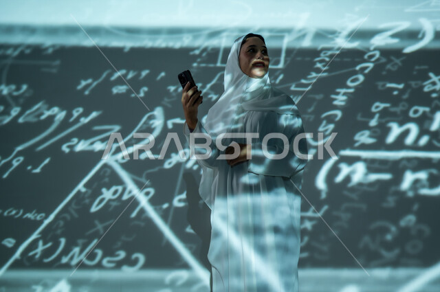 أرقام وحسابات في مادة الرياضيات، استخدام التقنيات الحديثة, دمج العلم بالتكنولوجيا، إمرأة عربية خليجية سعودية مبتسمة محجبة ترتدي العباءة تمسك بيدها الهاتف المحمول, التطور العلمي و التقني في المملكة العربية السعودية, خلفية سوداء للقوانين والمعادلات الرياضية الملونة