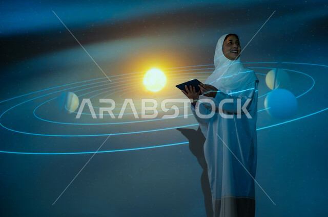 مسارات حركة الكواكب حول الشمس المتوهجة، استخدام التقنيات الحديثة، امرأة عربية خليجية سعودي مبتسمة محجبة ترتدي العباءة تقف أمام الكواكب والنجوم تمسك جهاز التابلت وتنظر الى شيء ما، الاهتمام بقطاع الفضاء في المملكة، خلفية هيولوجرامية ثلاثية الأبعاد