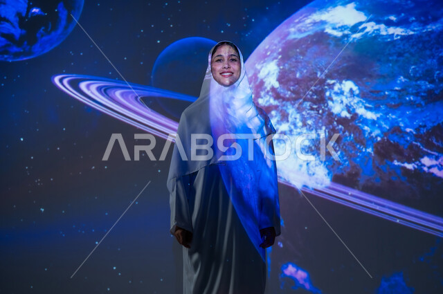 الاهتمام بقطاع الفضاء في المملكة، امرأة عربية خليجية سعودية مبتسمة محجبة ترتدي العباءة تقف أمام الكواكب والنجوم تنظر الى الكاميرا بإيماءات السعادة والسرور، تعزيز مفهوم التطور الرقمي والتكنولوجي، خلفية هيولوجرامية ثلاثية الأبعاد