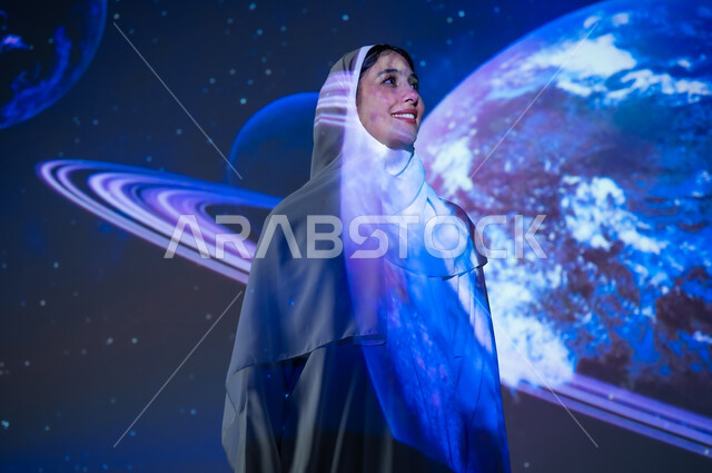 Promoting the concept of digital and technological development, a smiling Saudi Gulf Arab woman, veiled and wearing an abaya, standing in front of the planets and stars looking at something, interest in the space sector in the Kingdom, a 3D holographic background