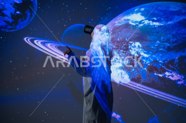 استخدام التقنيات الحديثة، امرأة عربية خليجية سعودية محجبة ترتدي العباءة تقف أمام الكواكب والنجوم تضع نظارات الواقع الافتراضي تشير بإصبع السبابة الى شيء ما، تعزيز مفهوم التطور الرقمي والتكنولوجي، الاهتمام بقطاع الفضاء في المملكة، خلفية هيولوجرامية ثلاثية الأبعاد