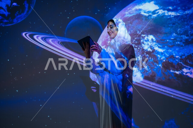 Promoting the concept of digital and technological development, interest in the space sector in the Kingdom, using advanced modern technologies, a smiling Saudi Arabian Gulf woman, veiled and wearing an abaya, standing in front of the planets and stars, holding a tablet and looking at it, a 3D holographic background