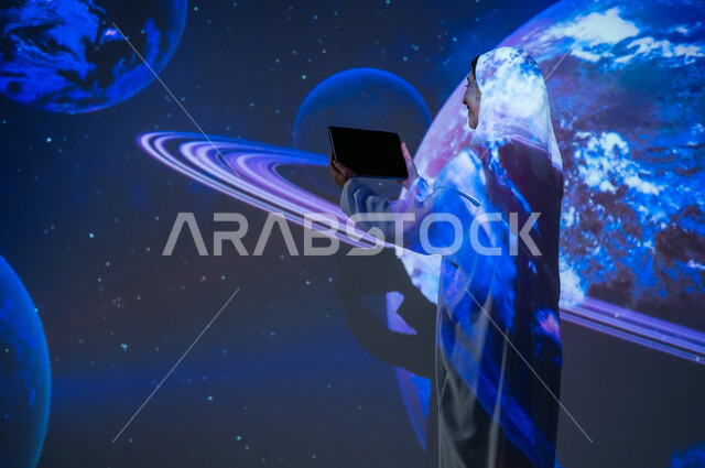 استخدام التقنيات المتطورة، صورة من الخلف لإمرأة عربية خليجية سعودية مبتسمة محجبة ترتدي العباءة تقف أمام الكواكب والنجوم تعرض شاشة سوداء فارغة عبر التابلت، الاهتمام بقطاع الفضاء في المملكة، تعزيز مفهوم التطور الرقمي والتكنولوجي، خلفية هيولوجرامية ثلاثية الأبعاد