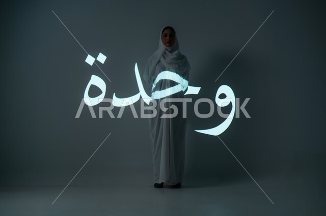Manuscript of the word unity in Arabic, portrait of a veiled Saudi Gulf Arab woman wearing an abaya looking at the camera with gestures indicating sadness, full-length body, gray background