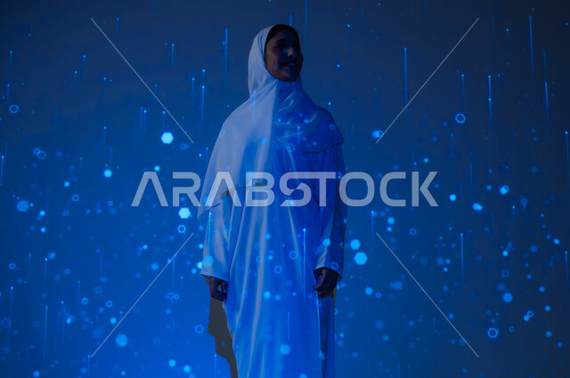 Promoting the concept of digital and technological development in Saudi society, a smiling Saudi Gulf Arab woman, veiled and wearing an abaya, looking at something, blue background with three-dimensional luminous holographic lines and circles