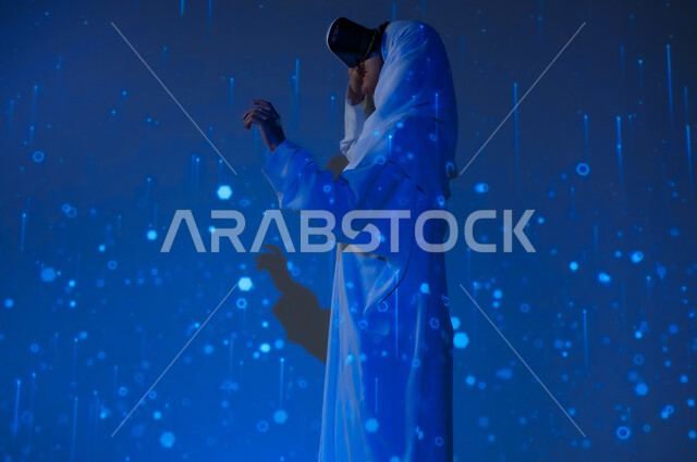 تعزيز مفهوم التطور الرقمي والتكنولوجي في المجتمع السعودي، استخدام التقنيات الحديثة، امرأة عربية خليجية سعودية مبتسمة محجبة ترتدي العباءة تضع نظارات الواقع الافتراضي وتشير بإصبع السبابة الى شيء ما، خلفية زرقاء مع خطوط ودوائر هولوجرامية مضيئة ثلاثية الأبعاد