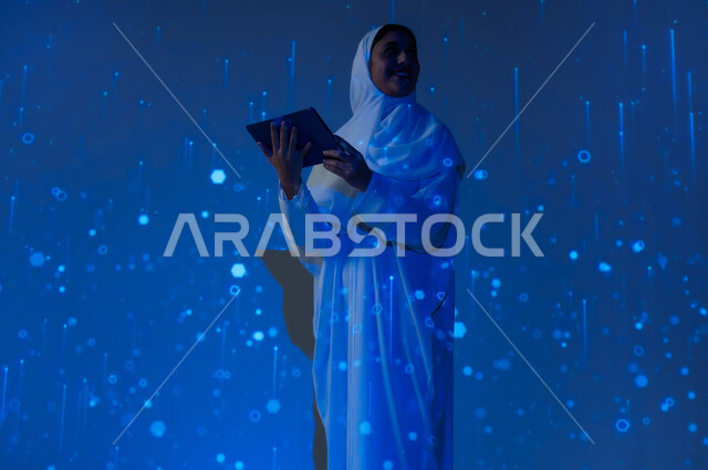 Promoting the concept of digital and technological development in Saudi society, using modern technologies, a smiling Saudi Arabian Gulf woman, veiled, wearing an abaya, holding a tablet and looking at something, a blue background with three-dimensional luminous holographic lines and circles