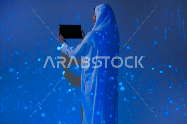 استخدام التقنيات الحديثة، تعزيز مفهوم التطور الرقمي والتكنولوجي في المجتمع السعودي، صورة من الخلف لإمرأة عربية خليجية سعودية مبتسمة محجبة ترتدي العباءة تعرض شاشة سوداء فارغة عبر التابلت، خلفية زرقاء مع خطوط ودوائر هولوجرامية مضيئة ثلاثية الأبعاد