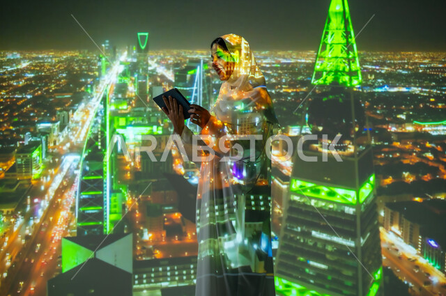 Using modern technologies, a smiling, veiled Saudi Arab woman wearing an abaya stands in front of King Fahd Road and Al Olaya Road in the city of Riyadh holding a tablet. The towers and skyscrapers are lit at night in green to celebrate National Day, September 23, and Saudi Flag Day, March 11.