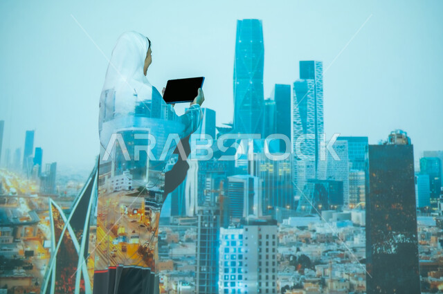 استخدام التقنيات الحديثة، صورة من الخلف لإمرأة عربية خليجية سعودية محجبة ترتدي العباءة تعرض شاشة سوداء فارغة عبر التابلت، المباني العالية وناطحات السحاب في مركز الملك عبد الله المالي ( كافد ) في الرياض, أماكن ومعالم مشهورة،  تطوير التصاميم الهندسية المعمارية