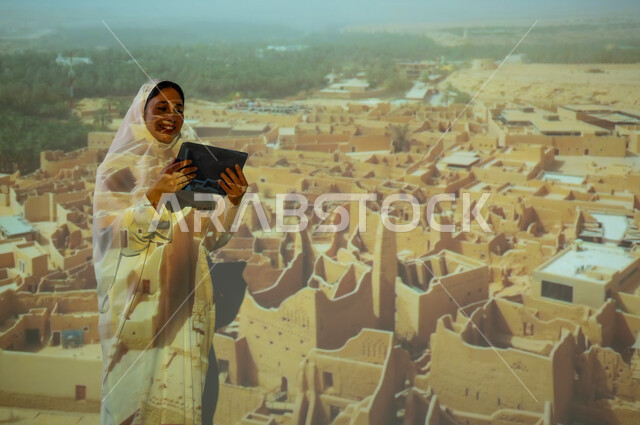 استخدام التكنولوجيا الحديثة والمتطورة، امرأة عربية خليجية سعودية مبتسمة محجبة ترتدي العباءة تمسك بيديها جهاز التابلت، عرض شاشة سوداء فارغة، المباني والبيوت الأثرية الطينية في الدرعية، جذب واستقطاب السائحين من مختف البلدان، أماكن ومعالم سياحية شعبية تراثية