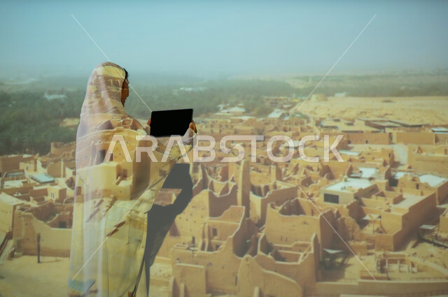 المباني والبيوت الأثرية الطينية في الدرعية، جذب واستقطاب السائحين من مختف البلدان، أماكن ومعالم سياحية شعبية تراثية، صورة من الخلف لإمرأة عربية خليجية سعودية محجبة ترتدي العباءة تمسك بيديها جهاز التابلت، استخدام التكنولوجيا الحديثة، عرض شاشة سوداء فارغة