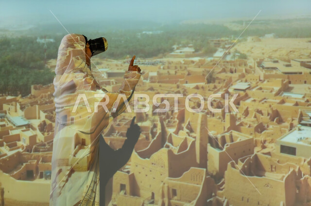 المباني والبيوت الأثرية الطينية في الدرعية، استخدام التقنيات الحديثة والمتطورة، صورة من الخلف لإمرأة عربية خليجية سعودية محجبة ترتدي العباءة تضع نظارة الواقع الافتراضي وتشير بإصبع السبابة لشيء ما، أماكن ومعالم سياحية شعبية تراثية، جذب واستقطاب السائحين