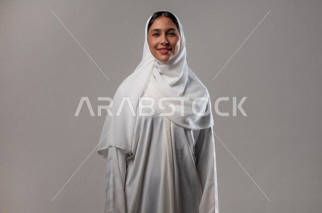 Taking personal photos for official documents, attention to elegance and external appearance, the concept of femininity and softness, a close-up portrait of a smiling Saudi Gulf Arab woman wearing a hijab and white abaya, looking at the camera with expressions of pleasure, gestures of self-confidence, gray background