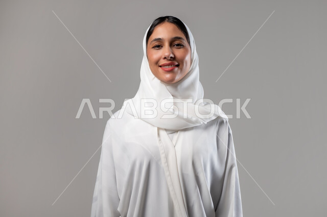 Concern with elegance and external appearance, the concept of femininity and softness, a close-up portrait of a smiling Saudi Gulf Arab woman wearing a hijab and white abaya, looking at the camera with expressions of pleasure, taking personal photos for official documents, gestures of self-confidence, gray background
