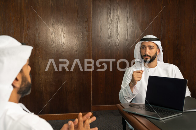 The concept of partnership and cooperation, using a laptop, two Arab Gulf Emirati employees in the meeting room inside the company headquarters, office jobs and professions, following up and discussing the company's strategies and objectives, studying and analyzing new projects