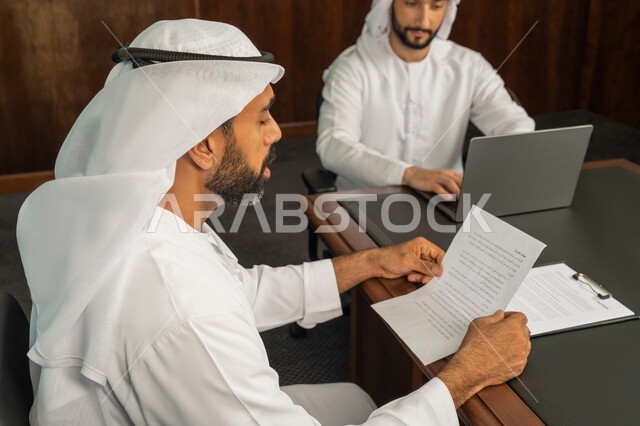الموافقة على الشروط وتوقيع العقود، رجل اعمال عربي خليجي اماراتي يرتدي الكندورة والغترة البيضاء يقوم بانجاز العمل عبر جهاز الحاسوب المحمول في الاجتماع، إبرام اتفاقيات وصفقات عمل ناجحة، مهن ووظائف مكتبية، الوحدة والعمل الجماعي