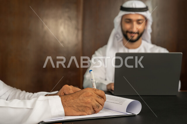الموافقة على الشروط وتوقيع العقود، رجل اعمال عربي خليجي اماراتي يرتدي الكندورة والغترة البيضاء يقوم بانجاز العمل عبر جهاز الحاسوب المحمول في الاجتماع، إبرام اتفاقيات وصفقات عمل ناجحة، مهن ووظائف مكتبية، الوحدة والعمل الجماعي