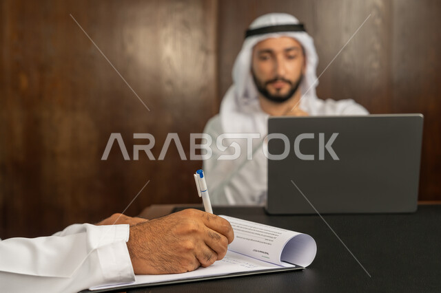 الوحدة والعمل الجماعي، الموافقة على الشروط وتوقيع العقود، رجل اعمال عربي خليجي اماراتي يرتدي الكندورة والغترة البيضاء يقوم بانجاز العمل عبر جهاز الحاسوب المحمول في الاجتماع، إبرام اتفاقيات وصفقات عمل ناجحة، مهن ووظائف مكتبية