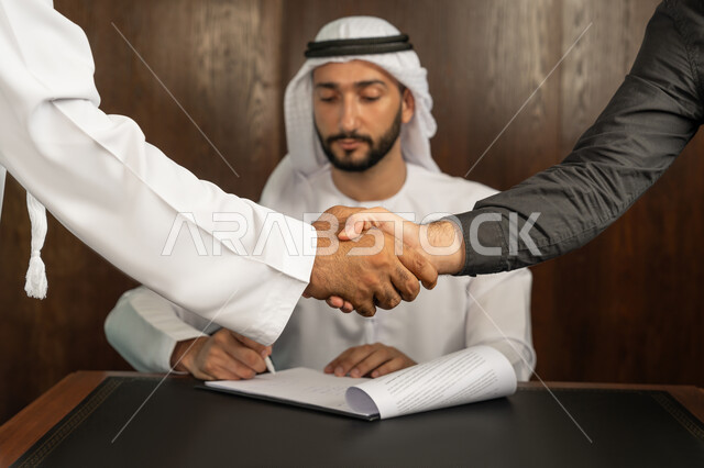 إبرام اتفاقيات وصفقات عمل ناجحة، مهن ووظائف مكتبية، الوحدة والعمل الجماعي، الموافقة على الشروط وتوقيع العقود، رجل اعمال عربي خليجي اماراتي يرتدي الكندورة والغترة البيضاء يقوم بانجاز العمل عبر جهاز الحاسوب المحمول في الاجتماع