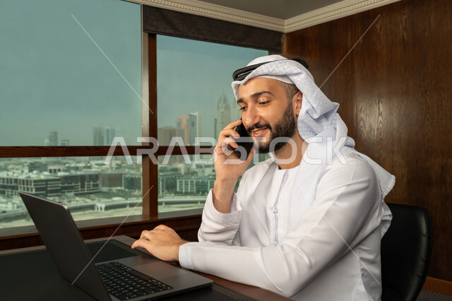 Completing tasks via laptop, integrating work with technology, using modern devices and technologies to complete office work, close-up portrait from the side of an Arab Gulf Emirati man wearing a kandura and ghutra making a phone call via mobile phone