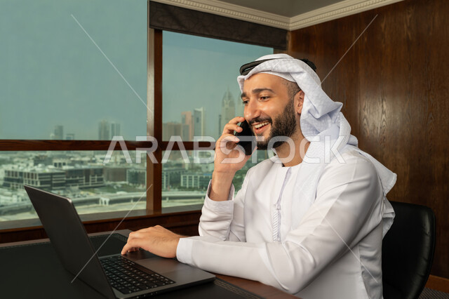 Integrating work with technology and technology, using modern devices and technologies to complete office work, close-up portrait from the side of an Arab Gulf Emirati man wearing a kandura and ghutra making a phone call via mobile phone, completing tasks via laptop