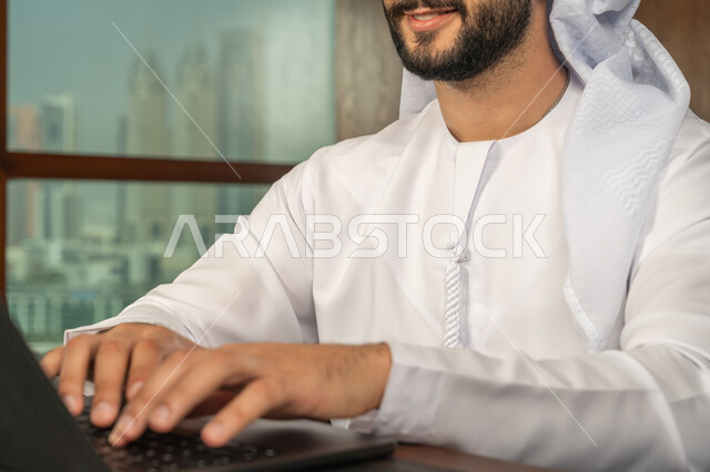 بيئة العمل المريحة في الشركات الاماراتية، مهن ووظائف ادارية مكتبية، مفهوم إدارة شؤون الأعمال وتنظيمها عبر الاجهزة التقنية، صورة مقربة لرجل أعمال عربي خليجي اماراتي مبتسم يرتدي الكندورة والغترة البيضاء يقوم باستخدام جهاز الحاسوب المحمول ضمن مقر الشركة