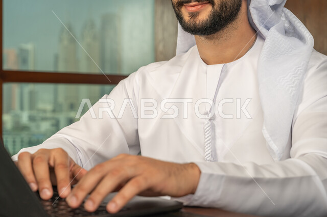 مهن ووظائف ادارية مكتبية، مفهوم إدارة شؤون الأعمال وتنظيمها عبر الاجهزة التقنية، صورة مقربة لرجل أعمال عربي خليجي اماراتي مبتسم يرتدي الكندورة والغترة البيضاء يقوم باستخدام جهاز الحاسوب المحمول ضمن مقر الشركة، بيئة العمل المريحة في الشركات الاماراتية
