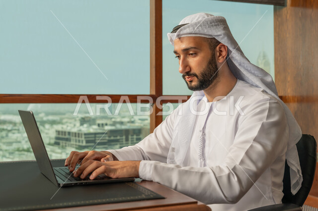 مفهوم إدارة شؤون الأعمال وتنظيمها عبر الاجهزة التقنية، مهن ووظائف ادارية مكتبية، صورة مقربة لرجل أعمال عربي خليجي اماراتي مبتسم يرتدي الكندورة والغترة البيضاء يقوم باستخدام جهاز الحاسوب المحمول ضمن مقر الشركة، بيئة العمل المريحة في الشركات الاماراتية