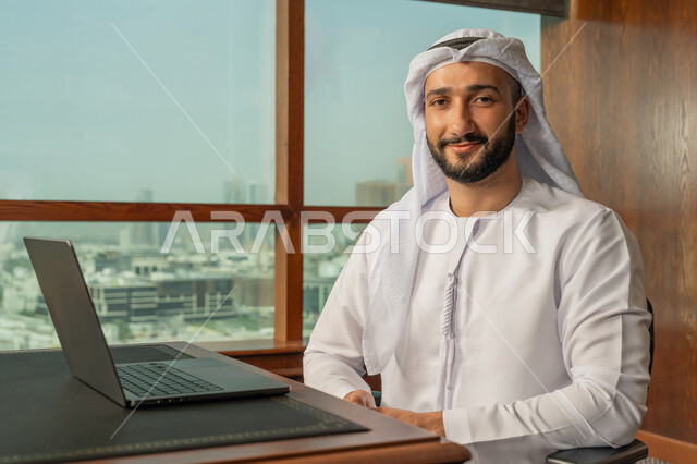النظر الى الكاميرا بتعابير السرور والسعادة، مهن ووظائف مكتبية، صورة مقربة لرجل أعمال عربي خليجي اماراتي مبتسم يرتدي الكندورة والغترة البيضاء يقوم باستخدام جهاز الحاسوب المحمول ضمن مقر الشركة، دمج العمل مع التقنية والتكنولوجيا