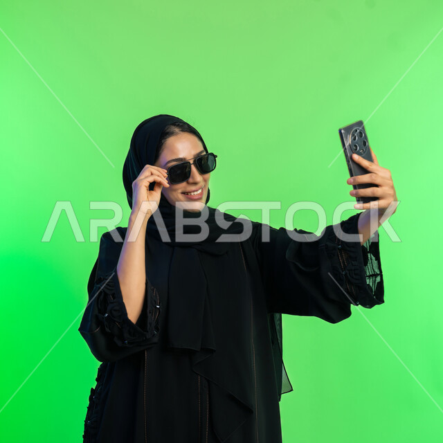 توثيق اللحظات السعيدة على وسائل التواصل الاجتماعي، النظر الى الموبايل بإيماءات السعادة، استخدام الأجهزة التقنية الحديثة، بورتريه لامرأة عربية خليجية سعودية محجبة ترتدي عباءة سوداء تلتقاط صور سيلفي تذكارية عبر الهاتف، شابة اماراتية أنيقة، خلفية خضراء، كروما