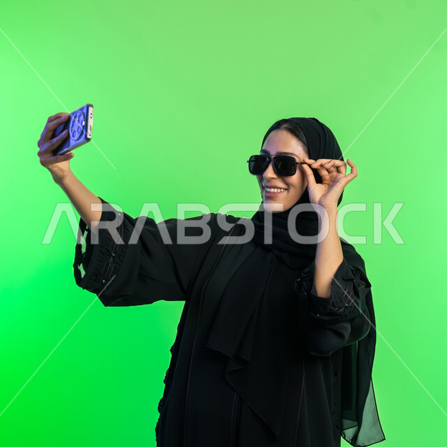 Looking at mobile with gestures of happiness, using modern technology devices, documenting happy moments on social media, portrait of a veiled Saudi Arabian Gulf woman wearing a black abaya taking souvenir selfies via phone, elegant Emirati young woman, green background, chroma key