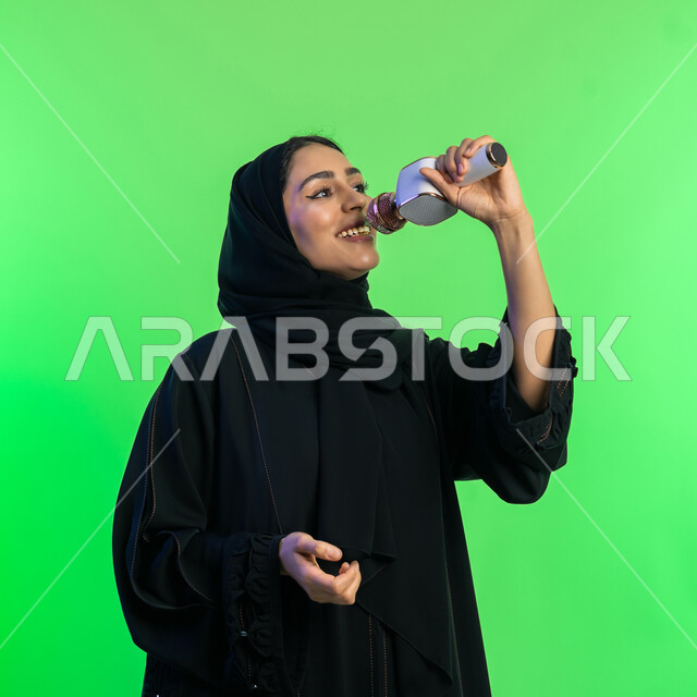 The concept of art and its positive impact on women, having a good time performing vocal performances, starting vocal training, close-up portrait of a veiled Saudi Arabian Gulf young woman wearing a black abaya holding a microphone, an Emirati woman practicing her singing talent with gestures of pleasure, green background, chroma key