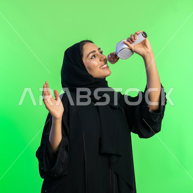 قضاء وقت ممتع بأداء العروض الصوتية، بدء التدريب الصوتي، مفهوم الفن وتأثيره الايجابي على الامرأة، بورتريه مقرب لشابة عربية خليجية سعودية محجبة ترتدي عباءة سوداء تمسك المايكروفون ، سيدة اماراتية تمارس موهبة الغناء بإيماءات السرور، خلفيه خضراء، كروما