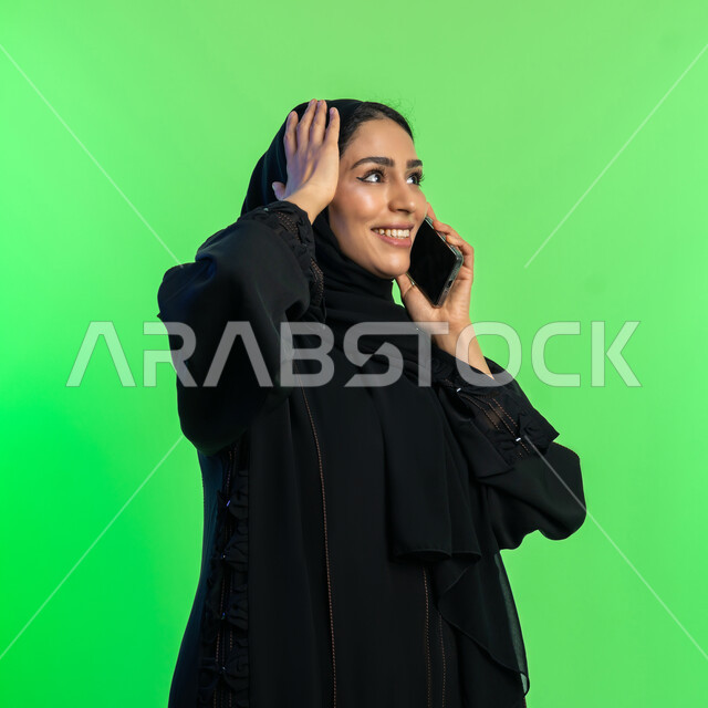 Gestures of integration and pleasure in communicating with relatives from a distance, making phone calls, close-up portrait of a veiled Saudi Arabian Gulf woman wearing a black abaya communicating with friends via mobile phone, young Emirati woman putting her hand on her head, green background, chroma key