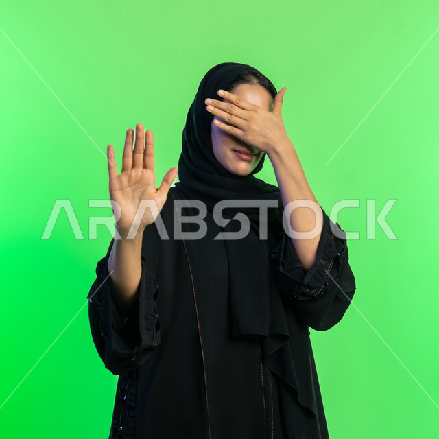 الأمر بالتوقف والابتعاد، الشعور بالإنزعاج وعدم الرغبة، شابة سعودية تغطي عينيها بتعابير الرفض والإمتناع عن رؤية شيء ما، بورتريه مقرب لامرأه عربيه خليجيه اماراتية محجة ترتدي عباءة سوداء ترفع يدها بإيماءات تدل على النهي، خلفية خضراء، كروما