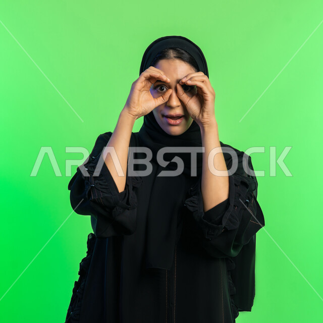 الأهداف والطموحات المستقبلية، حب الاستكشاف والمغامرة، بورتريه مقرب لإمراة عربية سعودية خليجية محجبة ترتدي العباءة السوداء تضع يديها على عينيها كالناظور، شابة اماراتية مبتسمة أنيقة، مفهوم تحديد الرؤية، خلفية خضراء، كروما