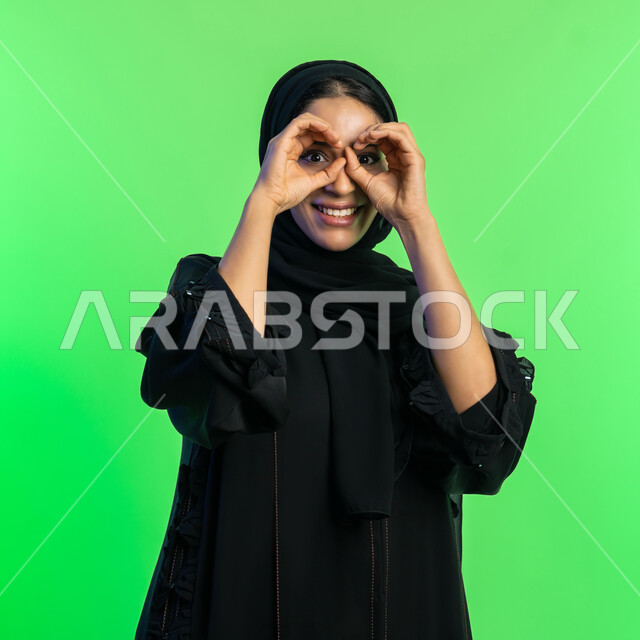 حب الاستكشاف والمغامرة، الأهداف والطموحات المستقبلية، بورتريه مقرب لإمراة عربية سعودية خليجية محجبة ترتدي العباءة السوداء تضع يديها على عينيها كالناظور، شابة اماراتية مبتسمة أنيقة، مفهوم تحديد الرؤية، خلفية خضراء، كروما