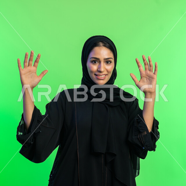 المفاجأة و الاندهاش من شيء ما، شابة سعودية تنظر الى الكاميرا بإستغراب، مفهوم النعومة والانوثة والاناقة، بورتريه مقرب لإمرأة عربية اماراتية خليجية مبتسمة محجبة ترتدي العباءة السوداء تقف باستقامة ترفع يديها نحو الأعلى بإيماءات الدهشة، خلفية خضراء، كروما