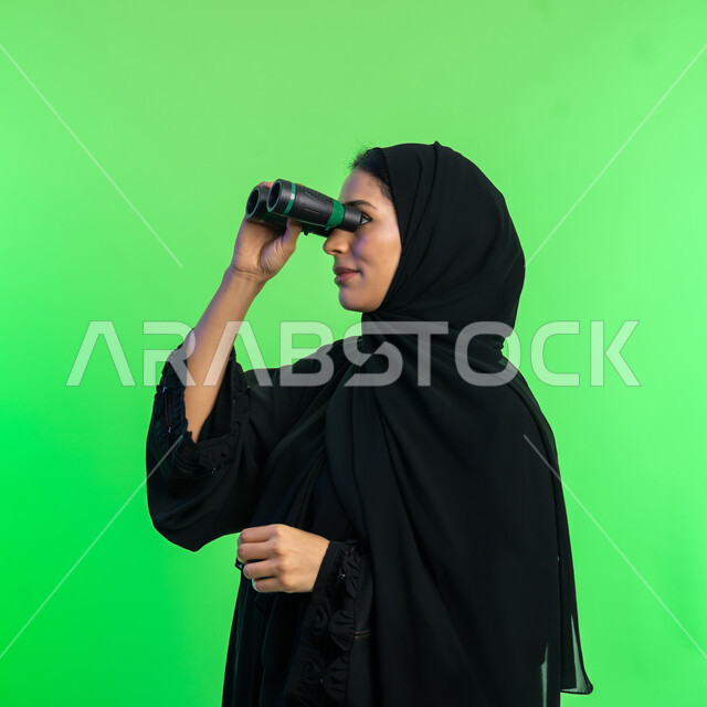 Seeing distant objects through a telescope, planning for future goals and ambitions, love of exploration and adventure, close-up portrait of a veiled Saudi Arabian Gulf woman wearing a black abaya looking through binoculars, elegant smiling young Emirati woman, green background, chroma key