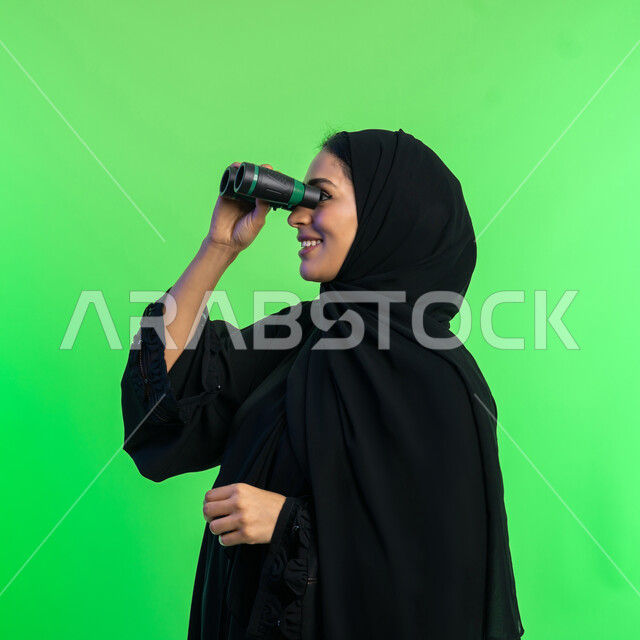 Future planning for goals and ambitions, love of exploration and adventure, seeing distant things through a telescope, close-up portrait of a veiled Saudi Arabian Gulf woman wearing a black abaya looking through binoculars, elegant smiling young Emirati woman, green background, chroma key